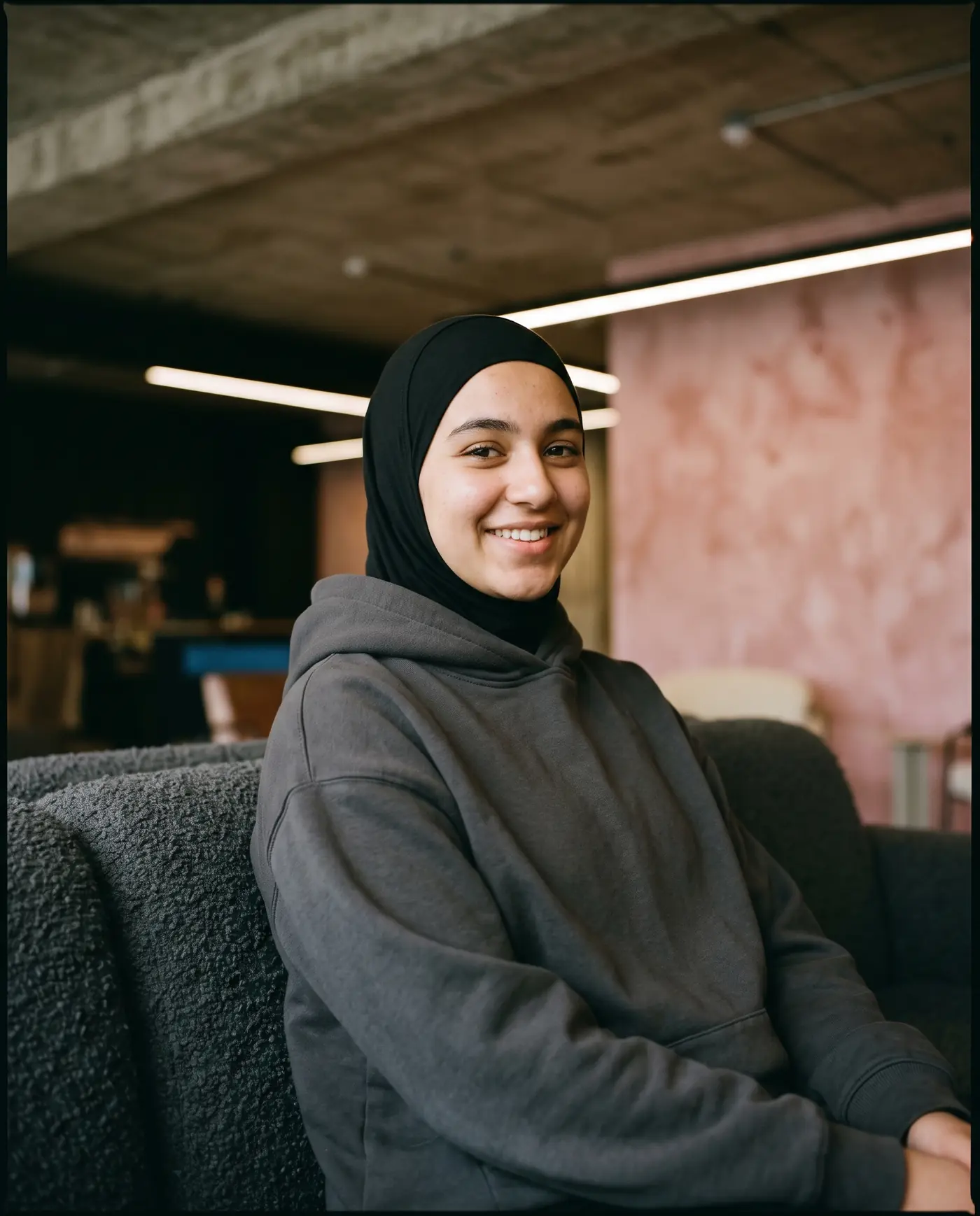 Porträtt av en leende tjej i sporthijab och grafitgrå hoodie i loungen.